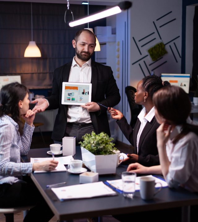 Business entrepreneur man presenting company statistics using tablet for financial presentation. Diverse multi-ethnic businesspeople brainstorming strategy working in office meeting room late at night